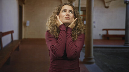 Fototapeta premium Young woman pressing hands to ears with eyes closed, curly hair, wearing a maroon sweater, near benches and stone columns in a building; discomfort.
