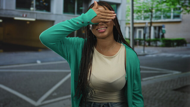 Woman in green cardigan covering eyes with hand and smiling on urban street near building crosswalk; shy joy.