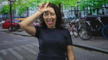 Fototapeta premium Woman makes l sign with hand to forehead and points finger toward camera on street with bikes and parked car; playful teasing.