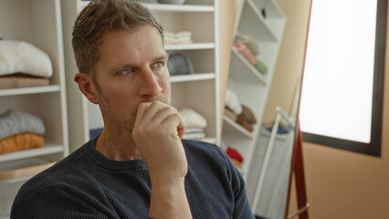Fototapeta premium Man hand to chin pensive in studio closet next to mirror and shelving with folded sweaters and knit navy jumper; contemplation solitude.