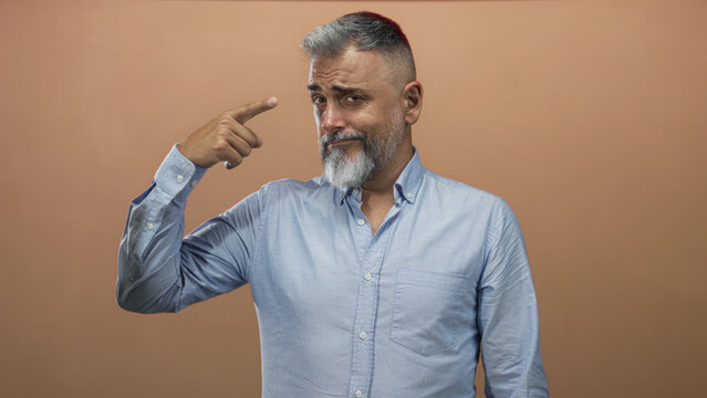 Man pointing finger to temple in studio with peach backdrop and light blue shirt, gray beard and raised eyebrow; wry humor skepticism.