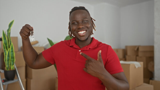 Man smiling and holding keys in hand points finger to keys inside a new house surrounded by moving boxes and potted plant; joy celebration.
