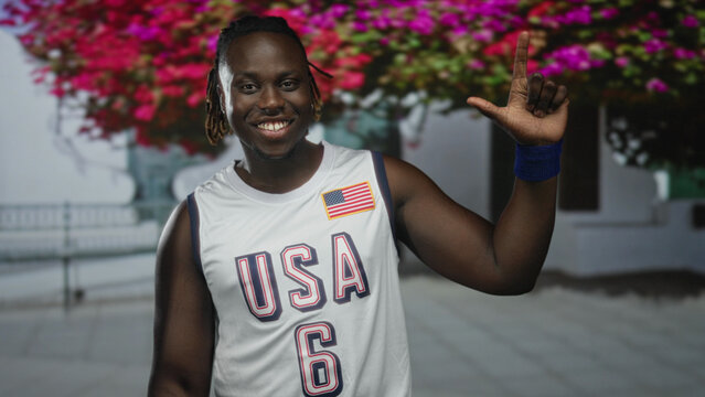 Young man in usa basketball jersey smiling and holding up peace sign with bare arm and blue wristband beside building and flowering trees; joy patriotism.