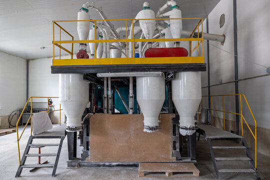 Industrial agricultural processing machinery with white cyclone separators and yellow safety railings stands inside a spacious facility for sorting and cleaning grains or other dry materials