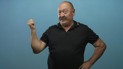 Naklejka premium Senior man smiling with clenched fist raised and hand on hip in studio against blue backdrop; confidence playfulness.