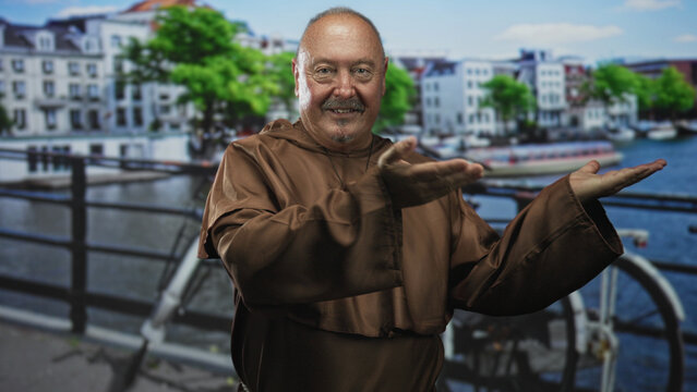 Man monk in brown habit with large cross necklace holds open palms toward camera on a street by a canal with railing and bicycle; welcome faith serenity.