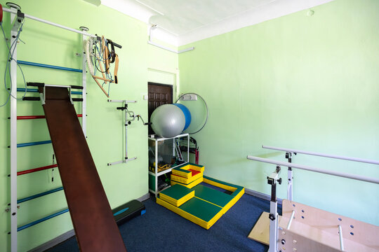 An empty physical therapy room with specialized exercise equipment including Swedish wall bars, a wooden slant board, parallel bars, balance balls, and soft foam blocks on a blue floor carpet