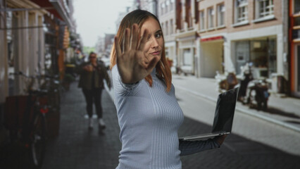 Fototapeta premium Woman holds laptop in one hand and extends palm forward in a crowded street, gaze stern and dismissive; defiance privacy.