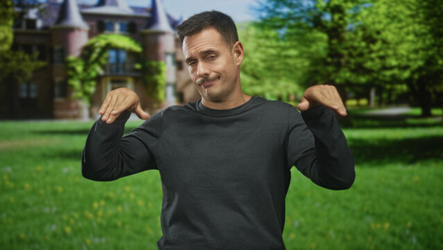 Man with moustache raises hands palms outward in front of a grand building, giving a rueful shrug; indifference.