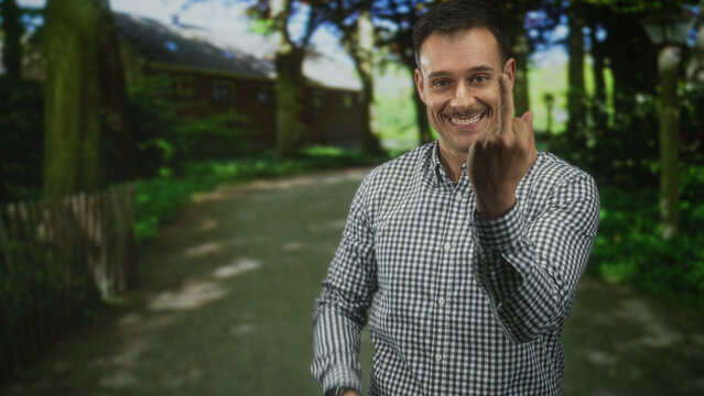 Man with moustache and checkered shirt showing middle finger and smiling while standing on a forest path near a cottage; defiance.