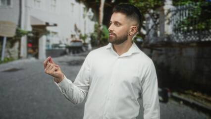 Fototapeta premium Hispanic man with beard and white shirt makes a pinching right hand gesture holding a small object while standing on a street; confidence.