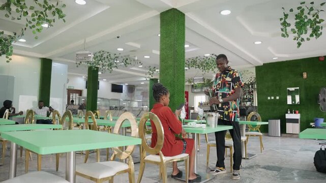 Black african couple romantic cafe date seated woman in red dress smiles while standing man offers small gift and lifts glass, patterned shirt contrasts lush plant covered interior, modern bright