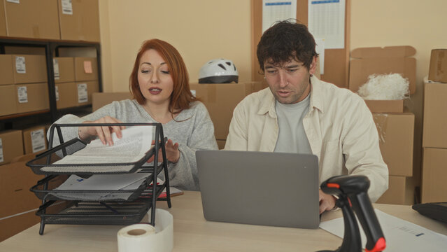 Man and woman point to a document and type on a laptop while sorting parcels and packing boxes in a building; focused teamwork.