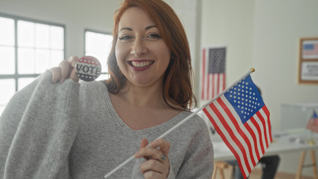 Woman holds vote button and waves american flag inside a building voting area beside a ballot box and voting booth; civic pride.