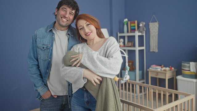 Couple cradling a swaddled baby in a home nursery with wooden crib, mobile and shelves; new parent joy.