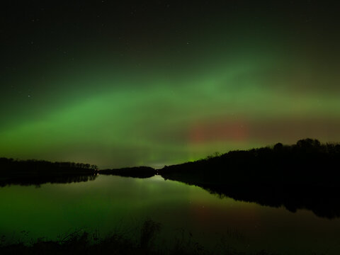 Northern lights of red and green fill the sky and reflect in the waters of a lake below