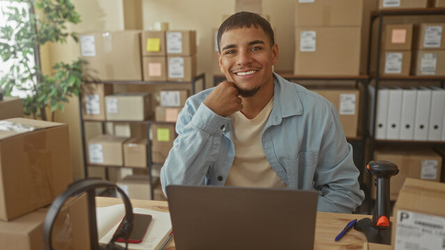 Man with hand on chin at desk with laptop and barcode scanner, surrounded by boxes and shelves in building; small business confidence.