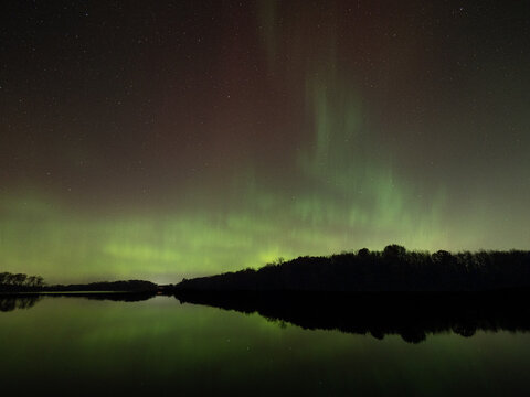Northern lights of red and green fill the sky and reflect in the waters of a lake below