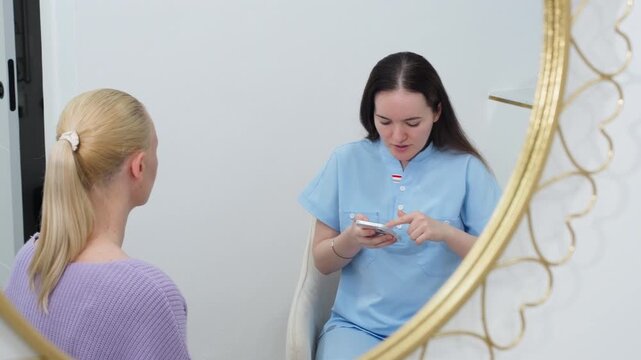 Doctor discussing personalized treatment plan with patient during consultation in modern cosmetology clinic, explaining aesthetic procedures and skincare options using smartphone reference.