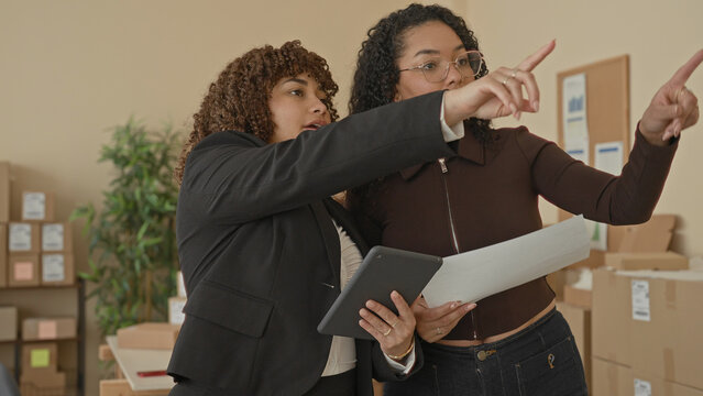 Woman holding paper and tablet, one points finger at stacked parcels and cardboard boxes in a building storage office; collaboration.