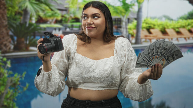 Woman holds dslr camera and fans us dollar bills in studio backdrop showing pool scene; confidence monetization business.
