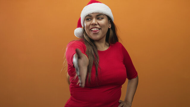 Woman extends right hand for handshake in studio wearing santa hat and red top while smiling and offering a greeting; playful welcome.