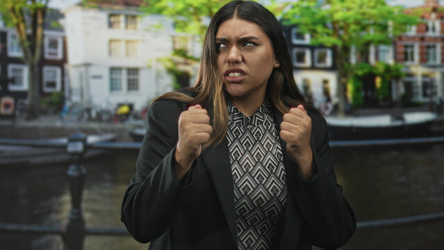 Woman grimaces and clenches fists on a canal street by residential buildings and a railing, face contorted and nose piercing visible; frustration resolve.