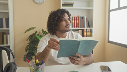 Man reading a paperback book, smiling and turning a page at a desk in a library building with bookshelves and a plant  serenity. © Krakenimages.com