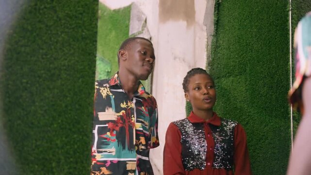 Young black couple posing by green wall, man leans close while woman smiles shyly, colorful patterned shirt, red dress with sequins, indoor portrait during date, tender romantic mood