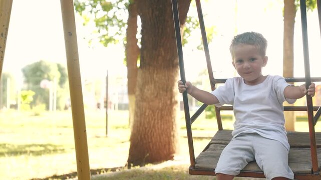 little boy swings on a swing. concept of a happy childhood and loving family. a child in white clothes swings on a swing on the lifestyle playground, the boy is being filmed by his parents