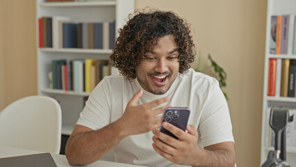 Man holding smartphone with thumb tapping screen while smiling at notifications, seated by bookshelves in a building  joy connection. © Krakenimages.com