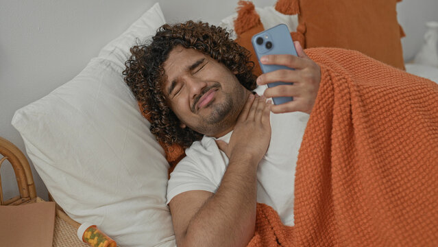 Young indian man lying in bed under orange blanket holds smartphone, touches throat with hand and winces beside pill bottle in studio; health concern.