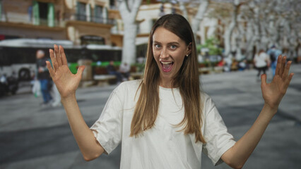 Fototapeta premium Woman with hands raised on street, palms open and smiling in a white tshirt with blurred bus and crowd behind her; joyful surprise.