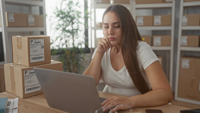 Woman typing on laptop beside stacked parcels processing online orders in office building; pensive small business planning.