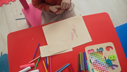 Naklejka premium Boy tracing a hand and holding a red crayon while coloring on paper at a red kids table in a studio with stickers and colored pencils; concentration.