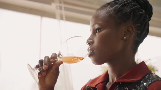 Young woman sipping wine at window, sequin top and braided hair, natural light highlighting amber beverage, reflective pause between sips as she waits for date, subtle smile with nervous anticipation,