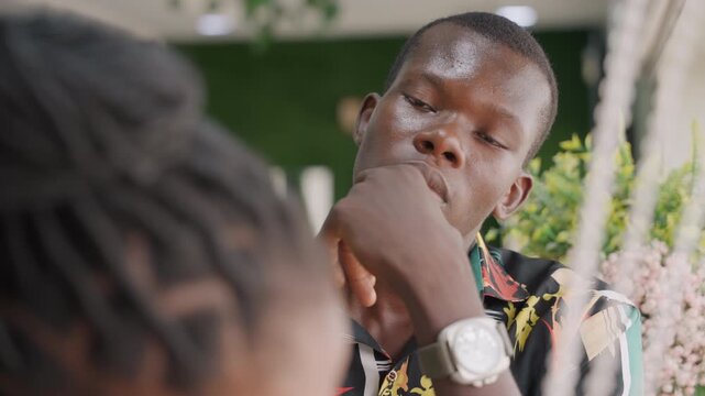Close candid portrait man touching chin, wristwatch visible, floral backdrop, thoughtful expression during date, rehearsing words, nervous pause, warm sunlight, street cafe ambience, subtle tension