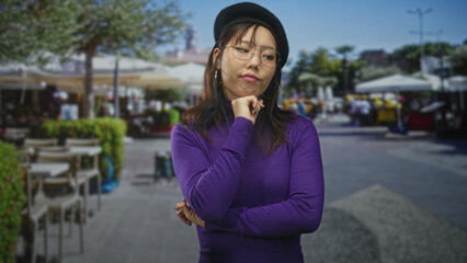 Fototapeta premium Young chinese woman in purple sweater with hand on chin and arms crossed, standing in a busy street cafe market plaza; thoughtful pondering doubt curiosity.