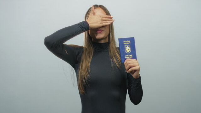 Woman holding ukrainian passport upright and covering her eyes with her hand in a studio with plain gray backdrop, passport emblem visible; shyness.