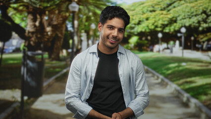 Fototapeta premium Man smiling with hands clasped at waist on a tree lined street path, wearing light shirt over black tee; warmth contentment.