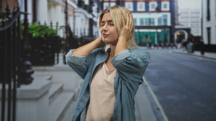 Fototapeta premium Woman covering ears with hands on a city street, denim shirt visible, wincing from headache; anxiety isolation.