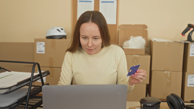 Woman holding creditcard and using laptop amid stacked packages at packing desk in building; focused small business owner.