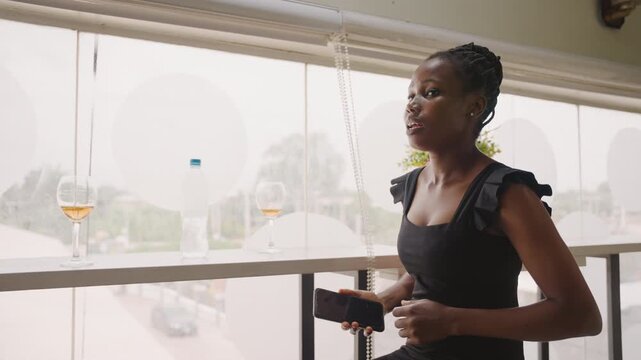 Black woman seated by window balcony looking out with soft smile, wine glass and flowers on sill, daylight city view, composing herself and rehearsing greeting, calm confident anticipation
