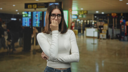 Fototapeta premium Young woman wearing glasses biting her nails with hand to mouth gesture in an airport terminal near departure screens and queues; travel anxiety delay.