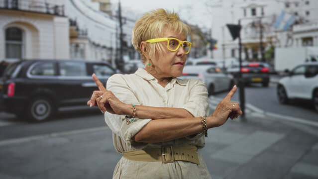 Woman senior hispanic with crossed arms and index fingers pointing outward, wearing yellow glasses and beige dress on a street near traffic and buildings; confidence assertiveness.
