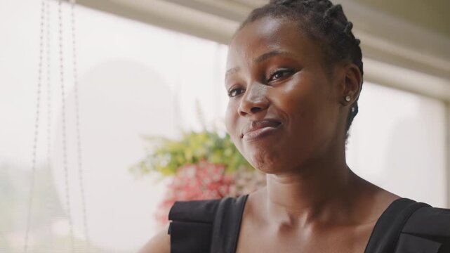 Black woman smiling by bright window, gentle profile closeup with braided hair, soft sunlight and floral backdrop, subtle playful expressions and reflective mood during cozy conversational date scene