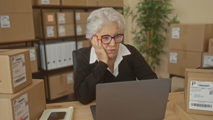 Fototapeta premium Elderly woman with laptop, hand on cheek, surrounded by shipping boxes, notebook and coffee on desk in building; concern resilience.