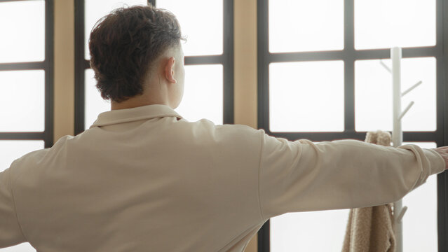 Young hispanic man with arms outstretched performing yoga t pose near large window in studio; serenity balance focus.