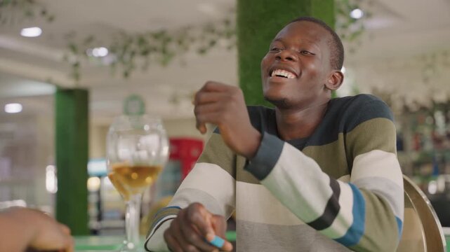 Smiling black man at cafe table engaged in playful conversation, holding phone, wine glass on table, greenery and warm string lights, striped sweater, flirty laughter and easy chemistry, intimate