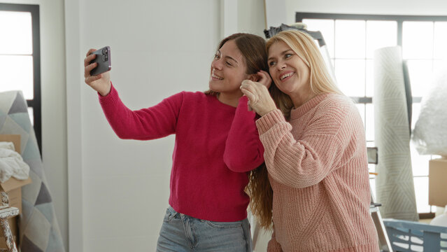 Woman taking selfie with smartphone, holding keys and smiling amid moving boxes in building; family bonding joy memories.